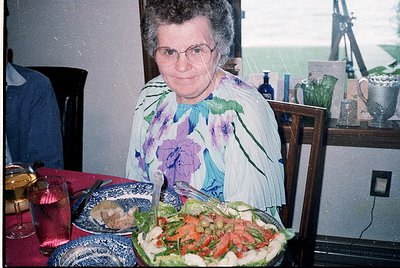 Vintage indoor portrait of a woman in a floral blouse, seated at a table with a large salad and blue-and-white dishware. Glas...