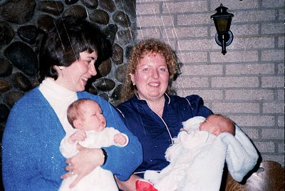 Two women holding infants in a cozy indoor setting with stone wall and vintage lamp. Likely 1980s–1990s family portrait. Warm...