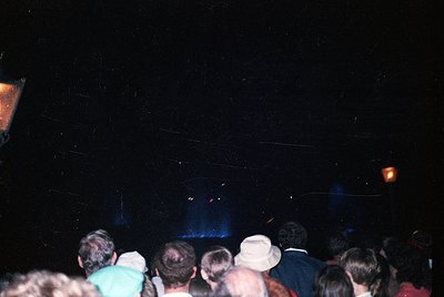 Vintage concert crowd shot, low-light stage lighting reveals blue haze. Casual 1990s attire—hoodies, caps, and jeans—suggests...