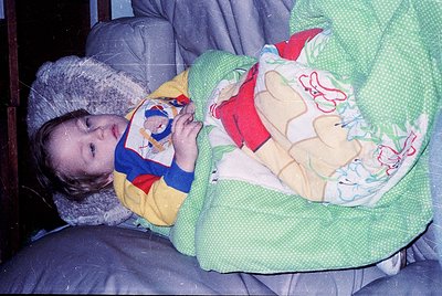 Child wrapped in vintage winter blanket with floral and cartoon snowman print, bundled in a knitted hat and yellow jacket. In...