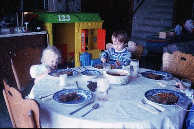 Vintage indoor scene: two young children seated at a round wooden table with ceramic plates of layered dessert, plastic cups,...