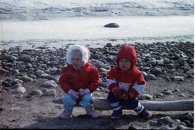 Two children in winter attire sit on rocky shoreline, playing in light snowfall. Both wear red jackets, hats, and mittens; on...