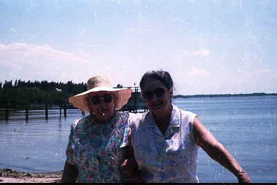 Two women pose by a waterfront in retro summer attire, likely mid-20th century. The woman on the left wears a wide-brimmed ha...