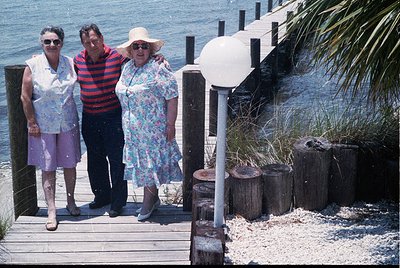Three adults pose on a wooden pier by the sea, 1970s-1980s. Woman left wears patterned blouse, purple pants, and sandals; man...