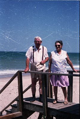Two elderly individuals pose on a wooden pier by the seaside, likely mid-20th century. The man wears a light-colored button-u...