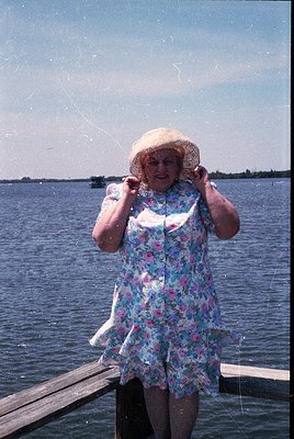 Vintage seaside portrait of a woman in a floral dress and straw hat, holding a fishing net near calm waters. Mid-20th century...