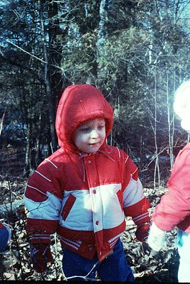 Vintage-style photo of a young child in a red-and-white winter jacket with a hood, standing in a wooded area. The jacket feat...