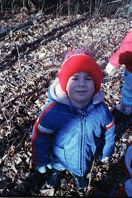 Child in winter attire—red knit hat with white trim, blue puffer jacket with red/orange stripes—playing in a wooded area cove...