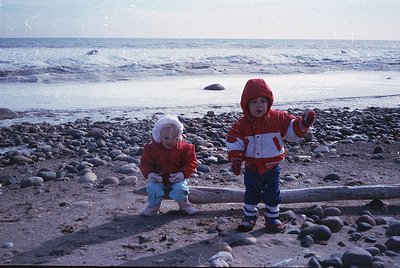 Two children in vintage winter gear explore a rocky seaside shore, 1970s-80s. Both wear red hooded jackets, white knit hats, ...