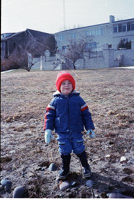 Child in winter gear—red knit hat, blue puffer jacket with red/white stripes, and black boots—standing on a grassy/sandy area...