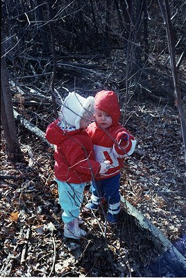 Two young children in matching red winter gear explore a wooded forest floor, likely late 20th century. The child on the left...