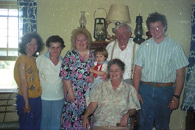 Family portrait from the 1970s–1980s, featuring seven individuals in a dimly lit indoor setting. Floral patterns dominate att...