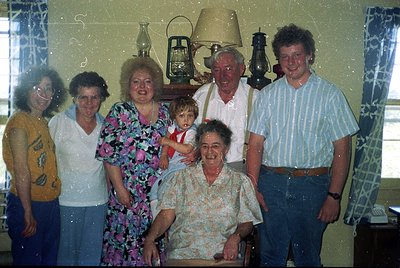 Family portrait from the 1980s–1990s featuring seven individuals in a dimly lit indoor setting. Central figure kneels holding...
