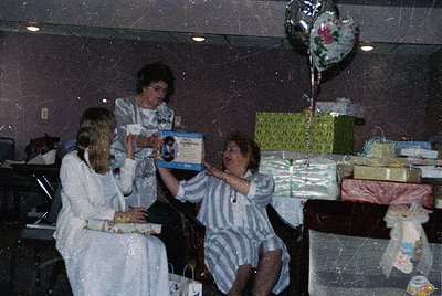 Vintage indoor celebration featuring three women in 1970s-style hospital gowns opening wrapped gifts. Decor includes a balloo...