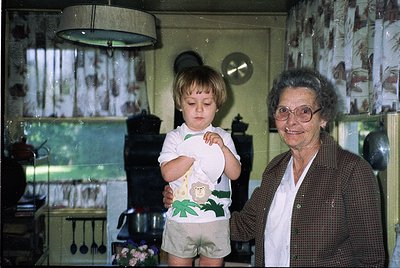 Vintage kitchen scene featuring a grandmother and toddler in mid-20th century home. Woman in patterned blazer poses beside ch...