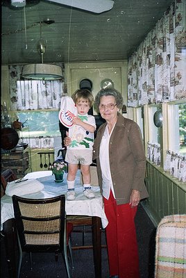 A grandmother and young child pose indoors in a modest, cluttered kitchen. The woman wears a patterned blazer, red trousers, ...