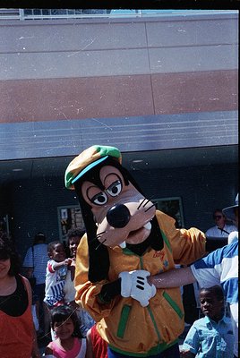 Goofy mascot in vintage Disney costume (yellow shirt, green hat) engaging with crowd at outdoor event. Reflective glass facad...