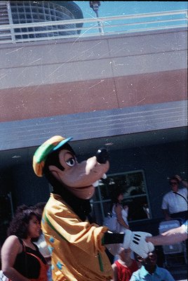 Mickey Mouse mascot in vintage costume (yellow shirt, green hat) interacting with crowd at outdoor event. Blurred background ...