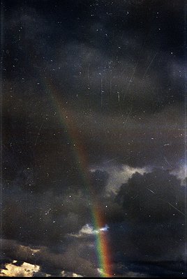 Vivid double rainbow piercing storm clouds under dramatic skies. Bold red/orange arc contrasts with softer blue/green. Raindr...