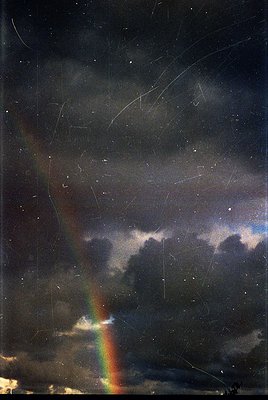 Vintage wet-plate or early film capture of a dramatic nighttime rainbow over storm clouds, with visible scratches and grain. ...