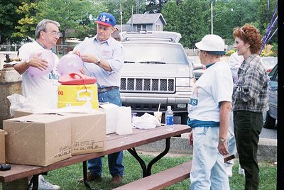 Community event setup in suburban setting, likely 1990s. Volunteers organize supplies: red cooler, yellow container, and card...
