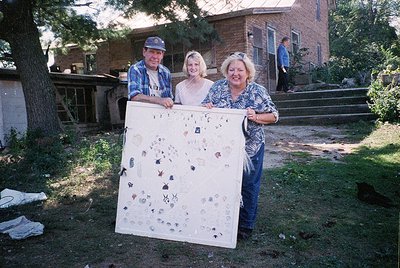 Three adults pose outdoors holding a large quilt adorned with appliquéd animals, flowers, and geometric shapes. The setting a...