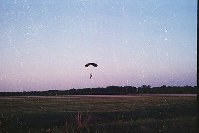 Vintage black-and-white photo of a parachutist descending over open fields at dusk, with dense forest in background. Film gra...