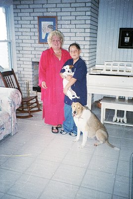 Two women pose indoors with dogs in a brick-walled room, likely mid-20th century. The woman in red wears a long robe; the wom...