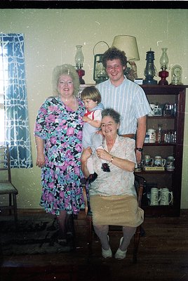 Vintage family portrait in a mid-century kitchen, featuring four generations: a woman in floral dress (left), a man in stripe...