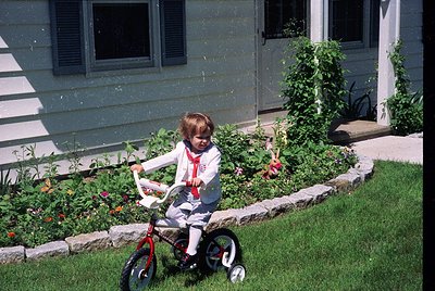 Young child in 1970s-style training wheels outfit rides red bicycle on lush suburban lawn. White vinyl siding house with clos...
