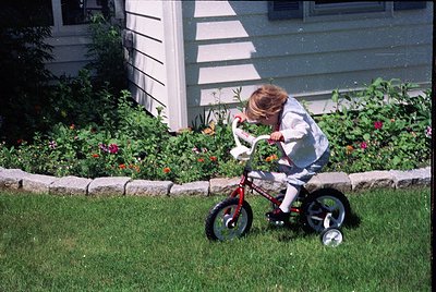 Young child on a red balance bike in a lush garden, surrounded by vibrant flowers and stone edging. White clapboard house in ...