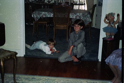 Vintage indoor scene featuring two children playing on a wooden floor in a mid-century dining area. The boy sits cross-legged...