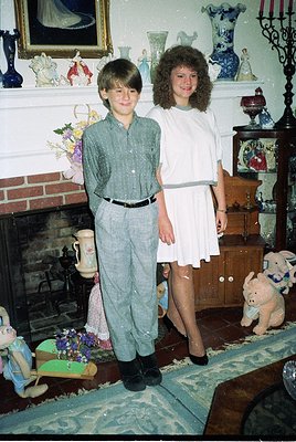 Two children pose indoors in a vintage-style living room, likely 1970s–1980s. Boy wears a button-down shirt, pleated trousers...