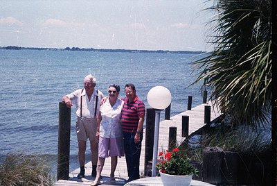 Three adults pose on a seaside wooden pier, 1970s–1980s coastal USA. Man in striped shirt, woman in floral dress, and man in ...