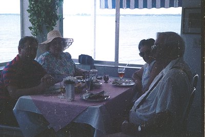 Four adults seated at a seaside restaurant table, enjoying a meal with drinks. Mid-century dining ambiance with red tableclot...