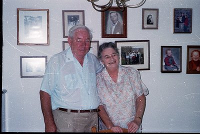 A 1980s/90s indoor portrait of an elderly couple posing in a home adorned with framed family photos. The man wears a light-co...