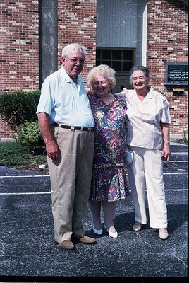 Three elderly individuals pose outdoors in front of a brick building with a plaque, likely mid-20th century (1960s–1970s). Th...