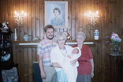 Family portrait in a rustic wooden interior, likely 1980s–1990s. Three adults and a newborn stand in front of a framed portra...