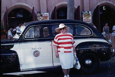 Vintage 1950s-60s police car with "POLICE" insignia parked near a retro-themed arcade. Woman in red-and-white striped dress a...