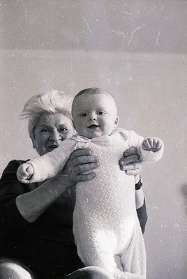 Vintage black-and-white portrait of an elderly woman holding an infant, both smiling directly at the camera. The woman’s hair...