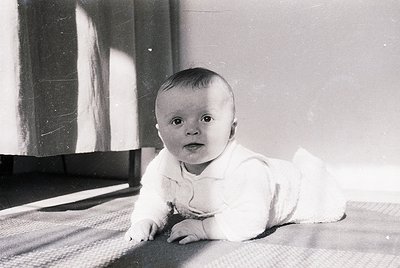 Vintage black-and-white portrait of an infant crawling on a patterned rug, dressed in a white onesie with a bow. Soft indoor ...