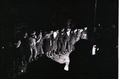 Black-and-white street scene capturing a group of men in formal attire (suits, ties, hats) standing in a line under dim light...