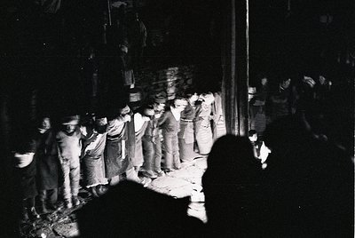 Crowd of men in 1960s-era suits and hats gathered outdoors at night, likely during a public event or demonstration. Motion bl...