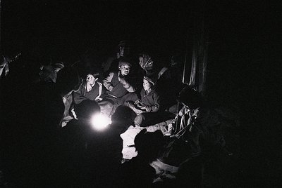 Black-and-white nighttime gathering in a dimly lit, enclosed space. Six individuals seated on wooden benches, illuminated by ...