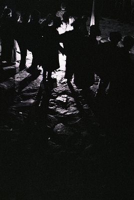 Silhouetted crowd dancing around a central fire pit on cobblestone ground, likely a traditional folk festival. Low lighting e...