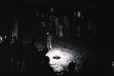 Black-and-white nighttime gathering in dimly lit courtyard, illuminated by single flashlight. Central figure in dark clothing...