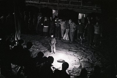 Black-and-white nighttime gathering in an open-air setting, likely a mine or industrial site. Central figure in white shirt a...