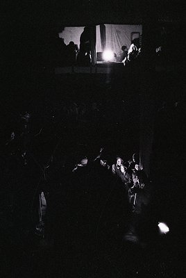 Low-angle shot of a dimly lit indoor crowd, silhouetted against a bright stage light. Central figures appear to be performing...