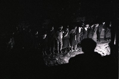 Black-and-white nighttime gathering in a dimly lit, enclosed space with a central fire. Group of men in traditional attire, p...