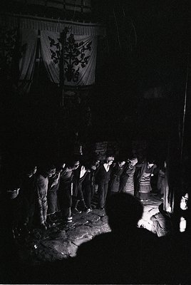 Group gathers around a central fire in a dimly lit courtyard, illuminated by flames. Embroidered banner with floral/foliage m...